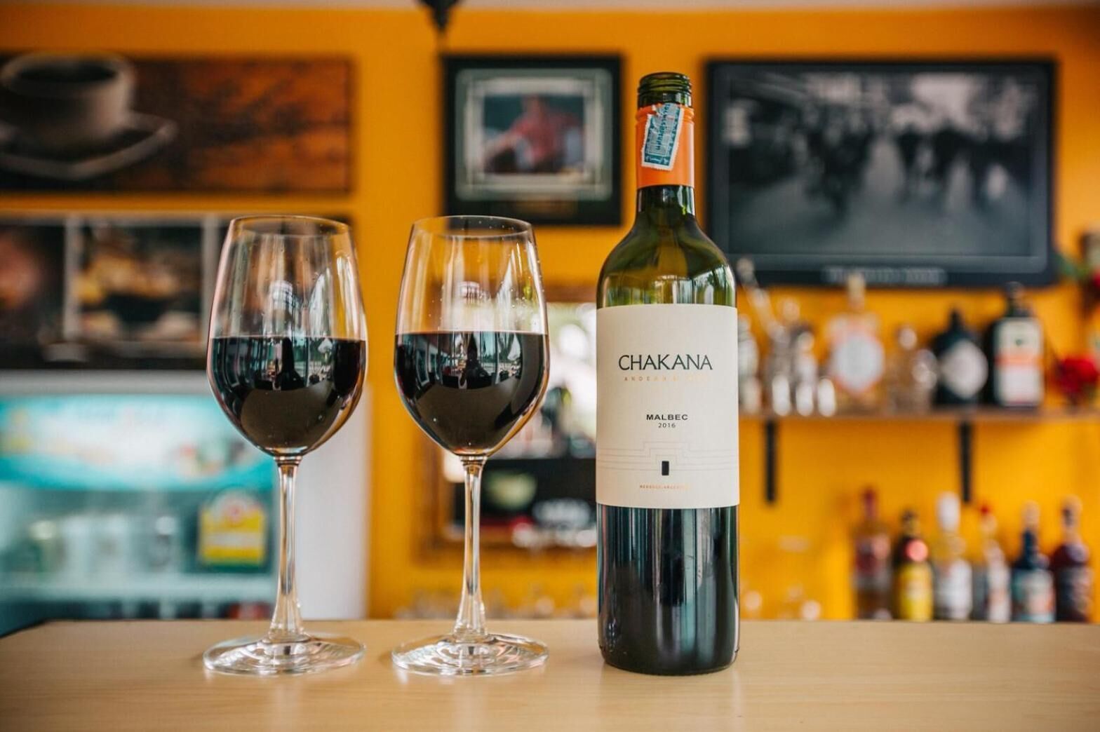 Dos copas de vino tinto con una botella de vino Chakan sobre una mesa en un bar con una pared amarilla y estantes llenos de botellas al fondo.
