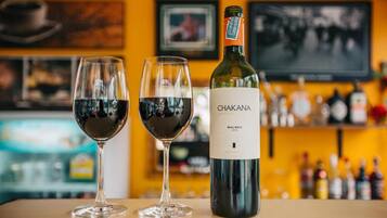 Dos copas de vino tinto con una botella de vino Chakan sobre una mesa en un bar con una pared amarilla y estantes llenos de botellas al fondo.