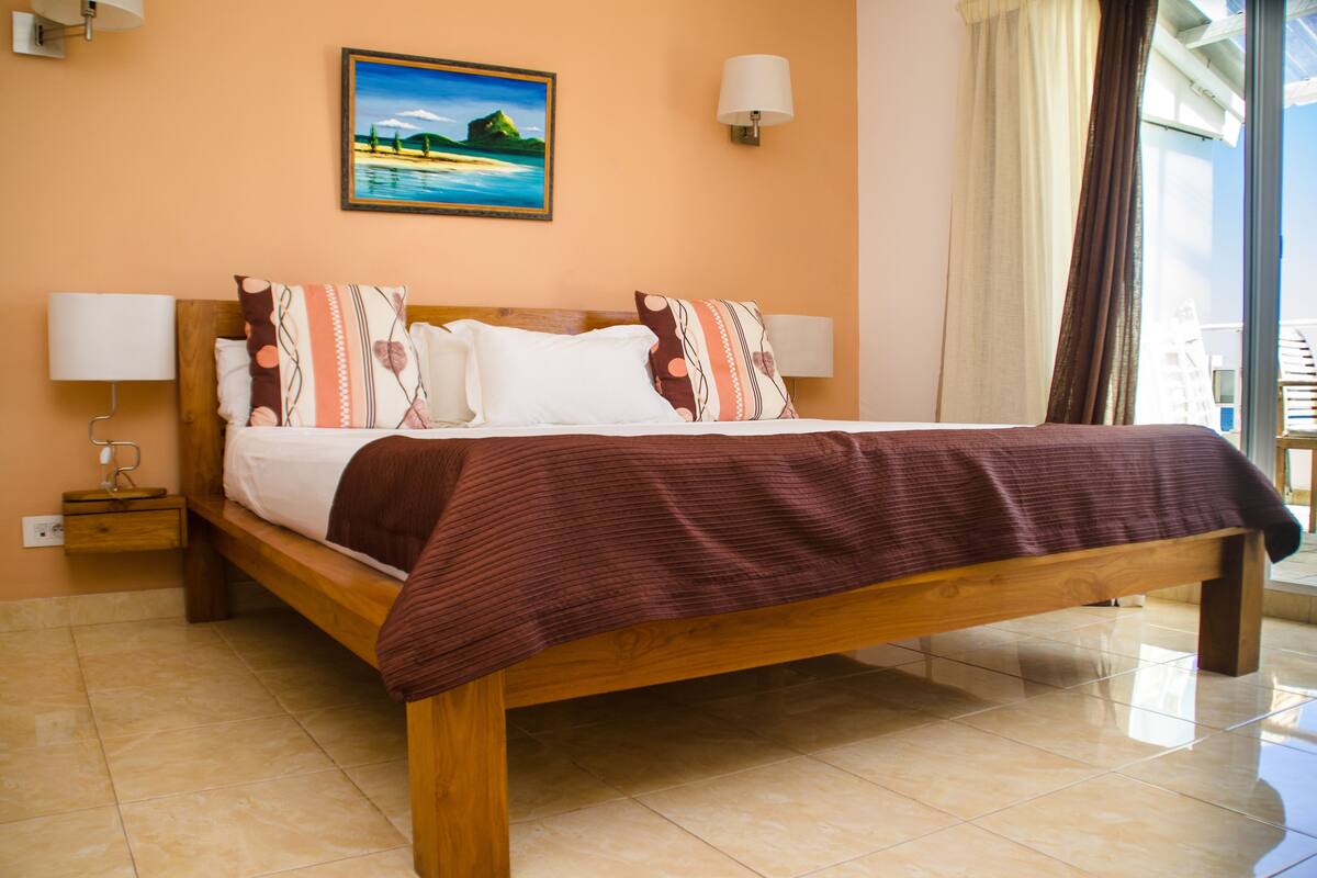 Un lit bien fait, avec un cadre en bois et une couverture marron, une table de chevet avec une lampe, et un tableau représentant une scène de plage accroché au mur.