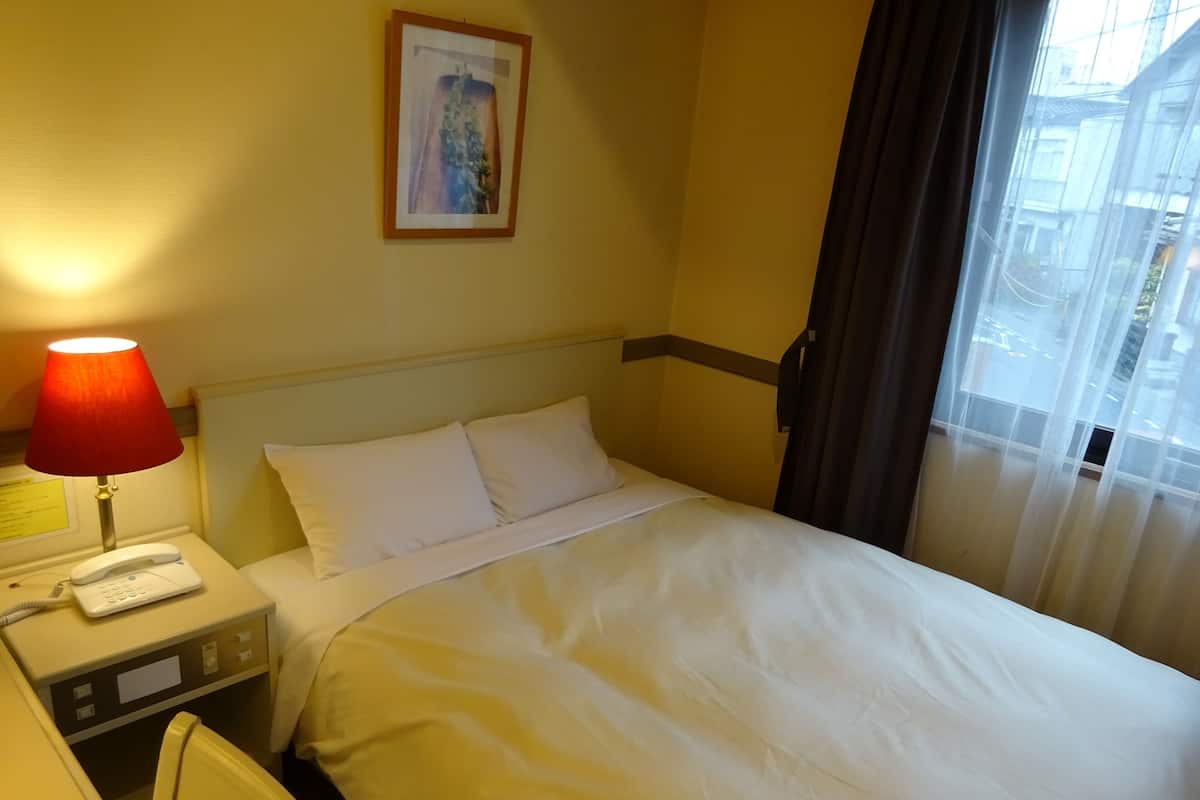 A hotel room with a bed, a desk with a telephone, a lamp, and a window with curtains.