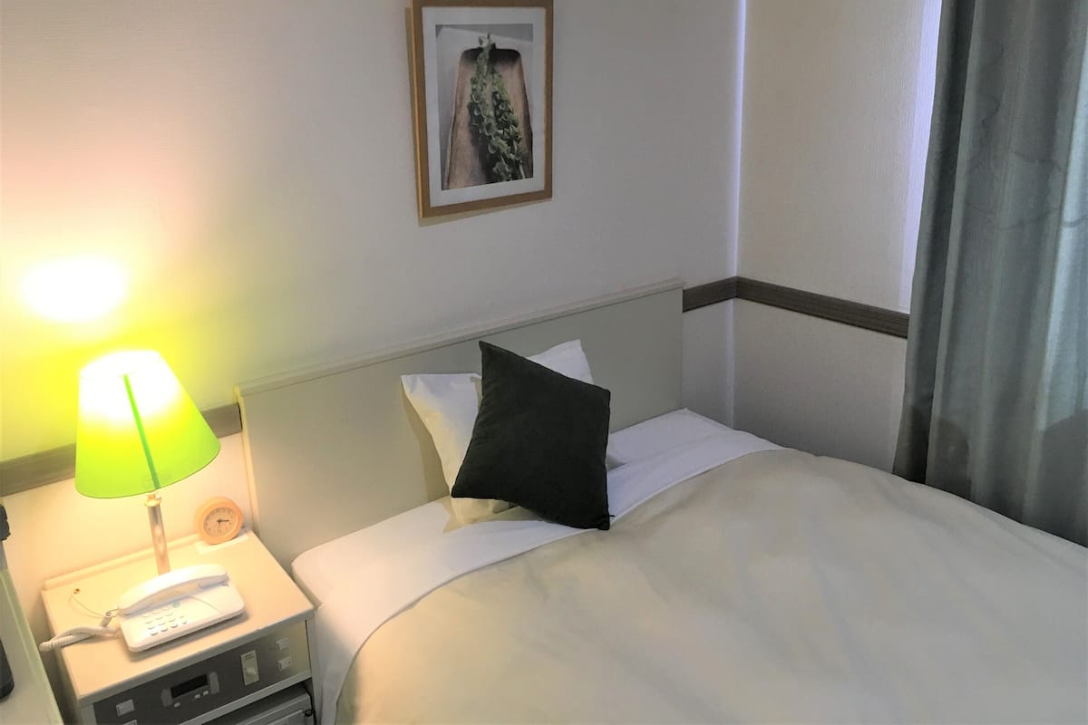 A hotel room with a bed, a nightstand with a lamp, a phone, and a clock.