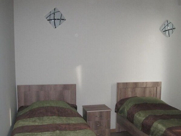 Une chambre avec deux lits, chacun doté d’une tête de lit en bois et d’une literie aux couleurs vertes et brunes. Une petite commode en bois se trouve entre les deux lits.