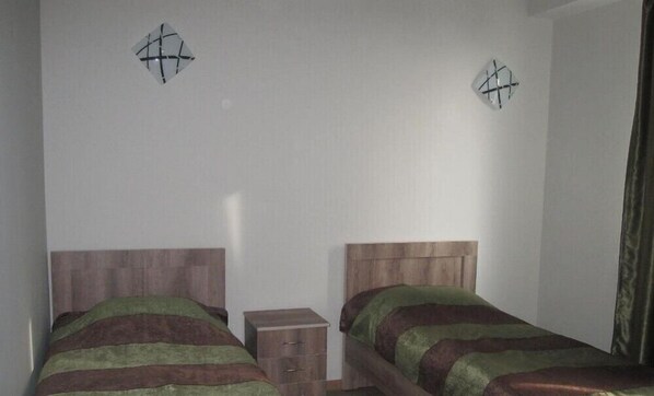 A room with two wooden beds, each with a green and brown striped bedspread. There are two small wooden nightstands with drawers, and the walls are plain white.
