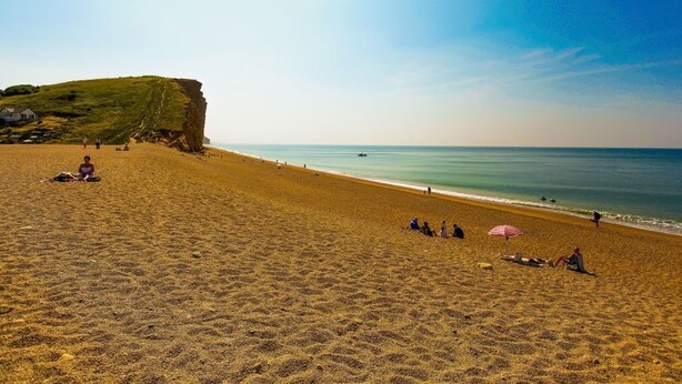 A beach with people sunbathing and walking along the shore, a cliff in the background, and a clear sky.