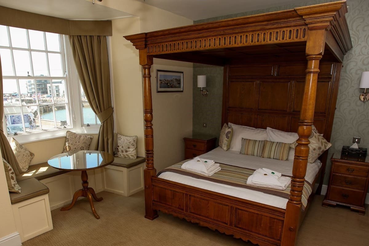 A four-poster bed with a wooden frame, a small round table, and a bench by the window.