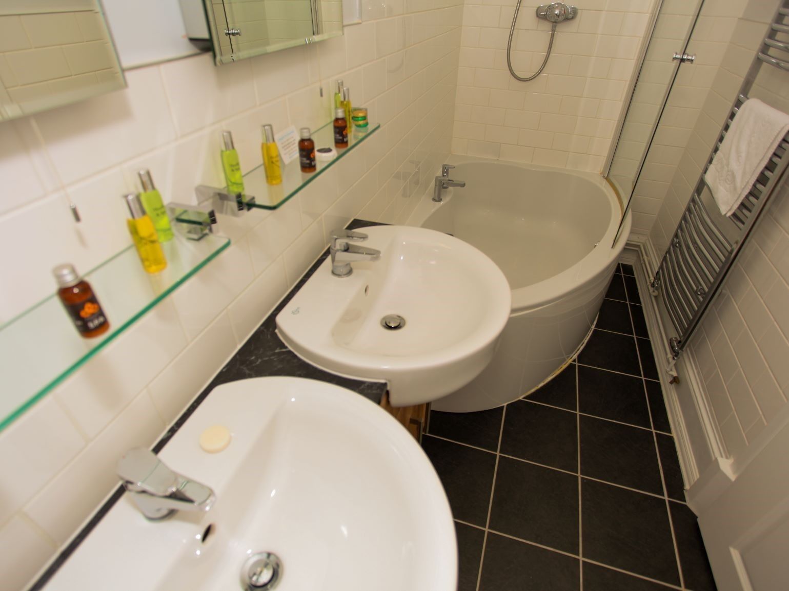 A bathroom with a bathtub, two sinks, and a glass shelf with toiletries.