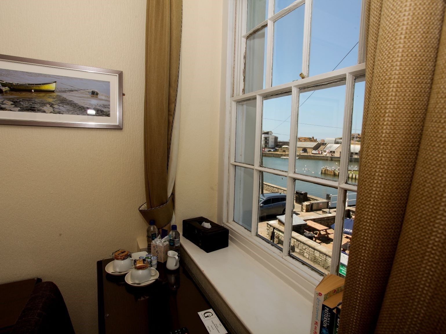 Ein Hotelzimmer mit Blick auf einen Hafen, ein kleiner Tisch mit einer Kanne und Tassen sowie ein gerahmtes Bild an der Wand.