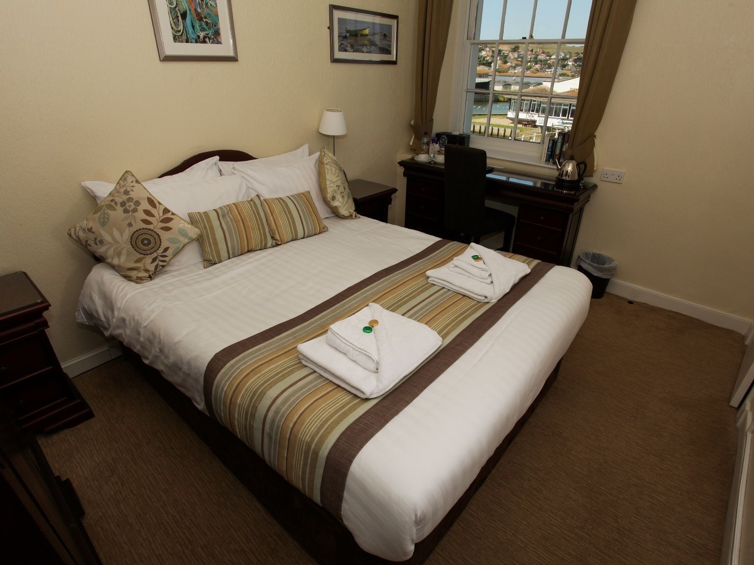 A neatly made bed with striped bedding and pillows, a bedside table with a lamp, and a view of a harbor through a window.