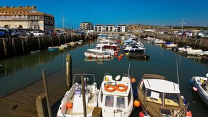 A marina with various boats docked, a wooden pier, and buildings in the background.