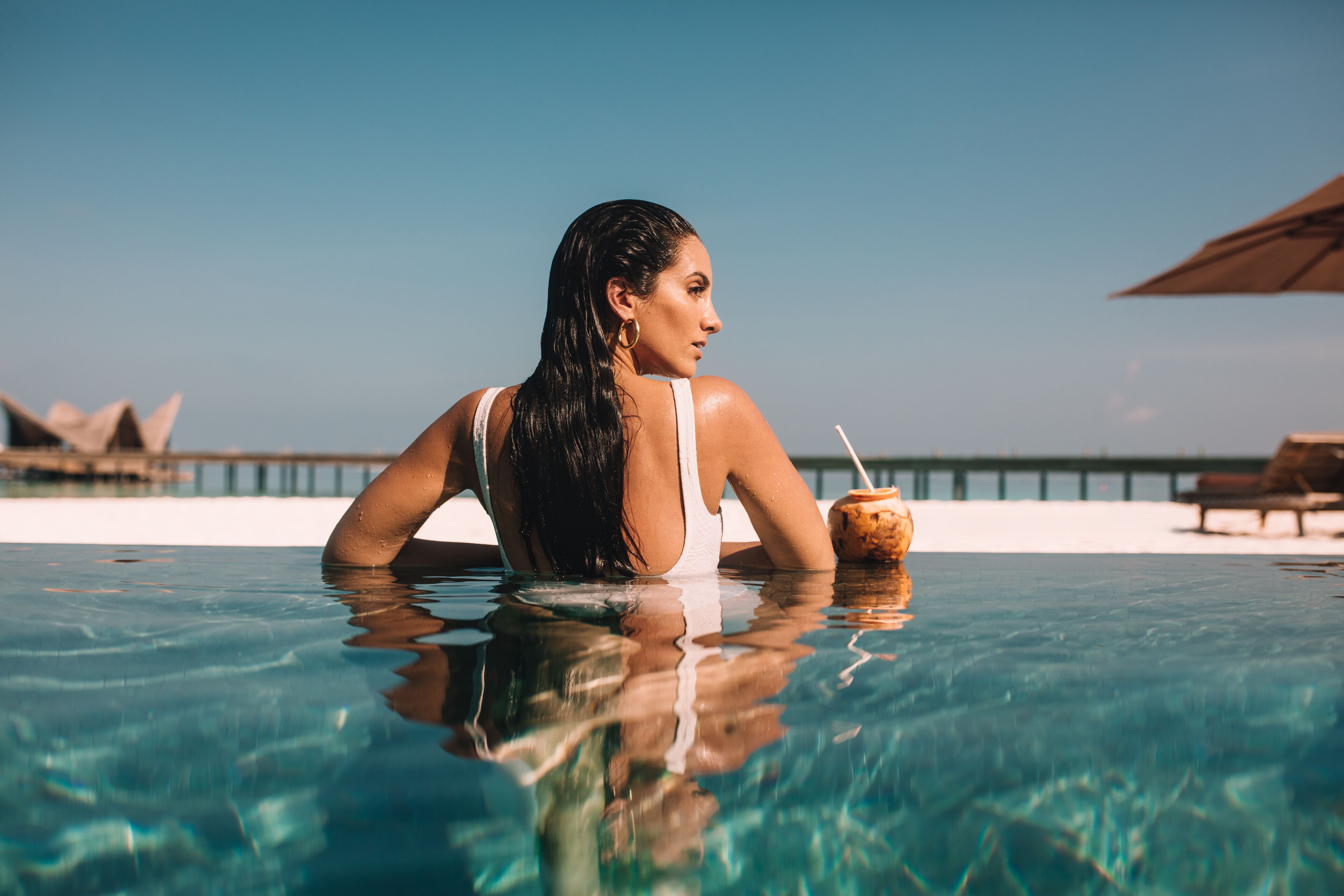 A person in a swimming pool with a drink, clear water, and a distant pier.