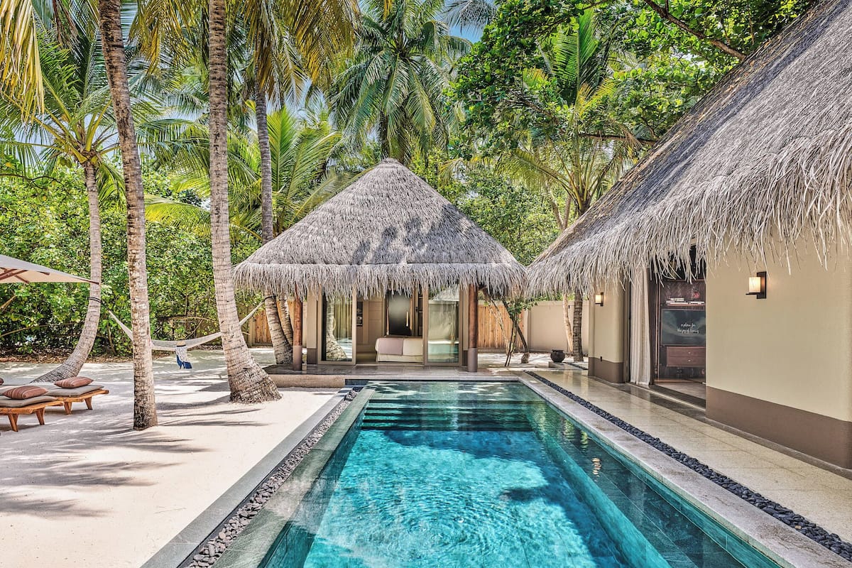 A pool area with a clear blue pool, surrounded by thatched-roof huts and palm trees.