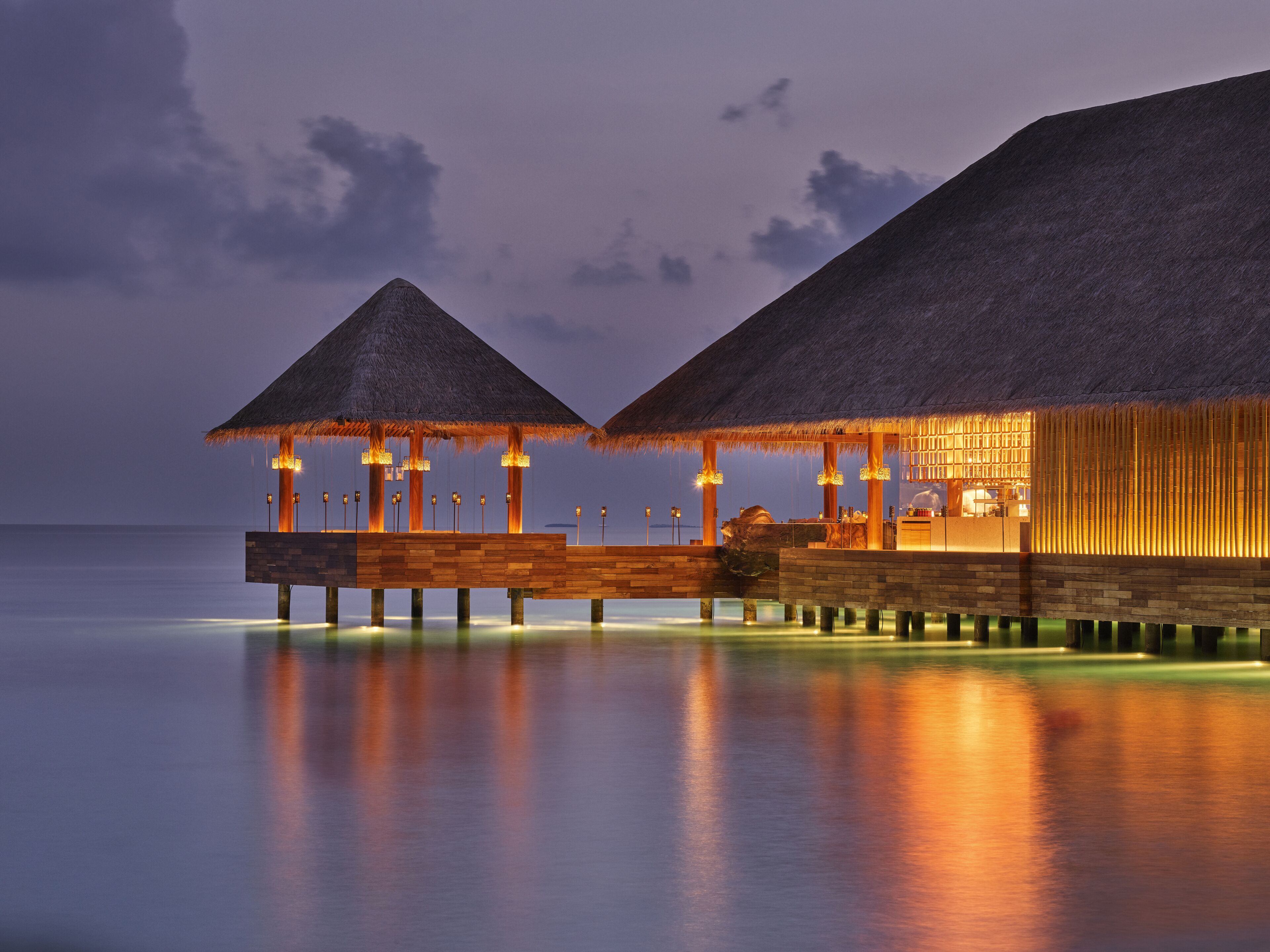 A thatched-roof overwater bungalow with a wooden deck, a bar area, and a view of the sea.