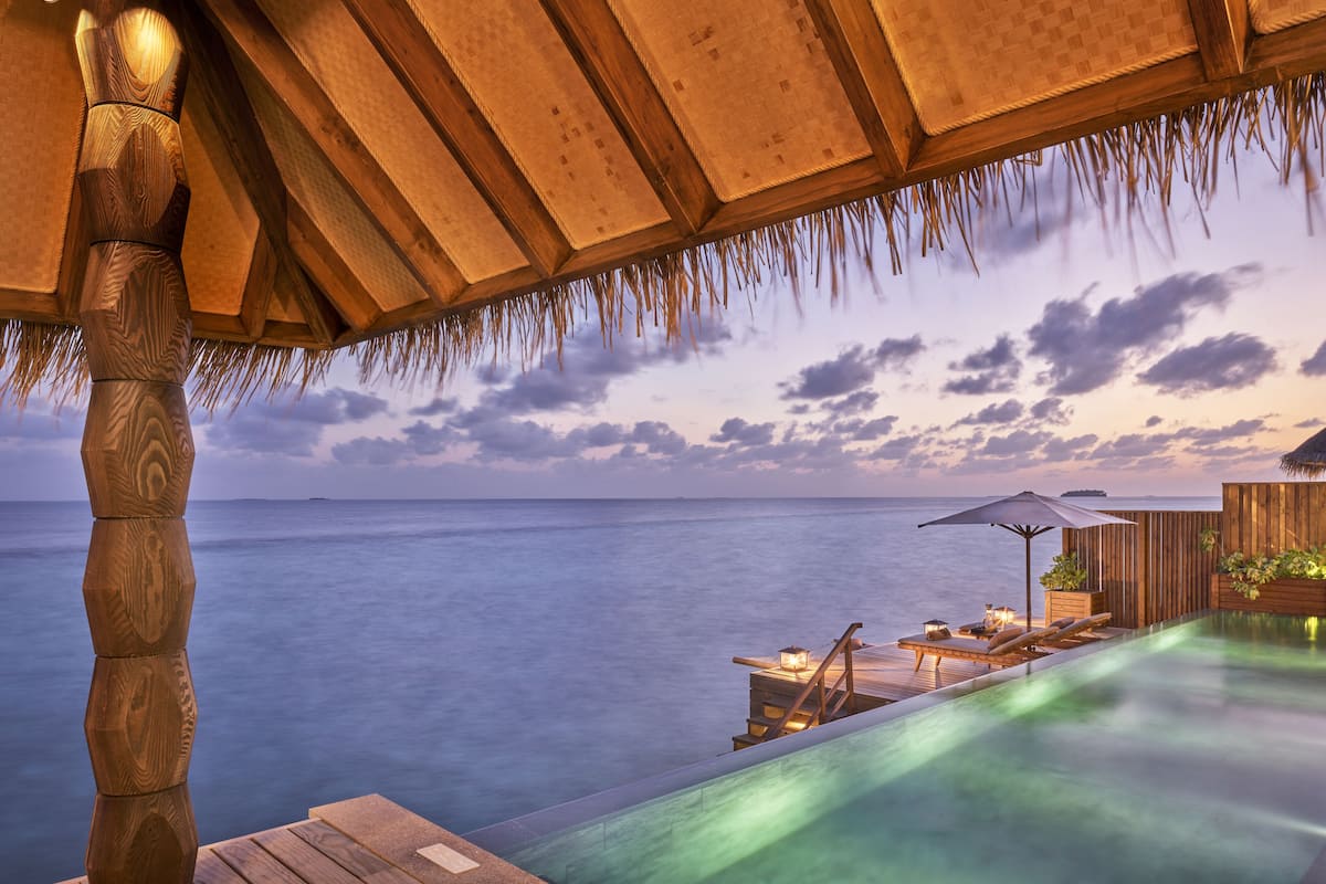 A wooden deck with a pool overlooking the sea, wooden furniture, and a thatched roof structure.