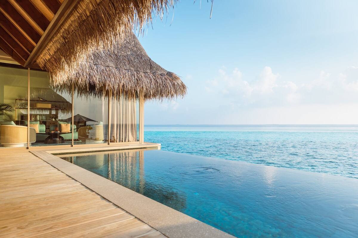 An infinity pool with a clear view of the ocean, a thatched roof structure, and a glass-enclosed room with furniture.