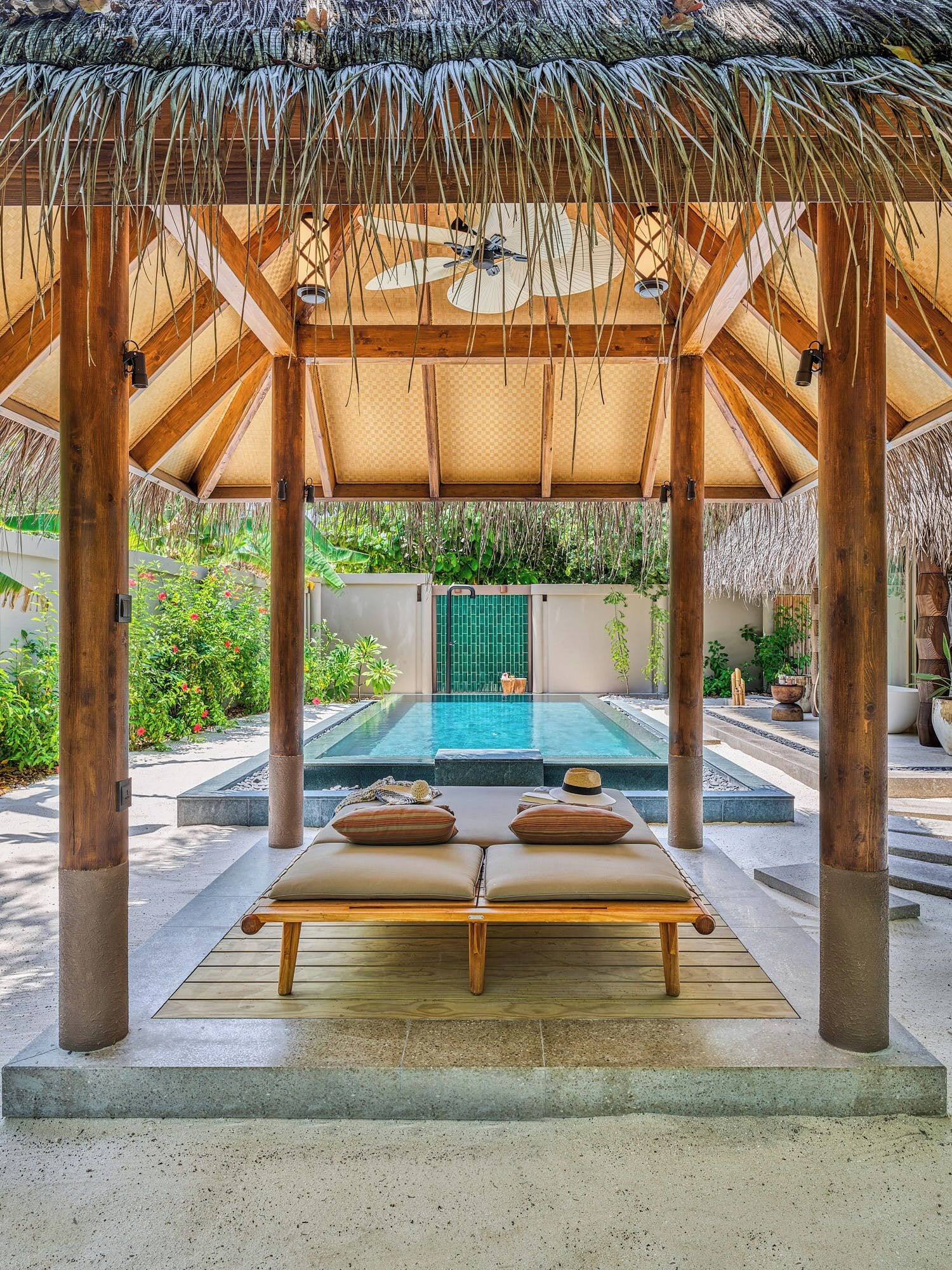 A covered outdoor area with a pool, two lounge chairs, and a thatched roof structure.