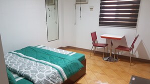 Une petite pièce propre, avec un lit, un bureau, deux chaises et une fenêtre avec des stores.