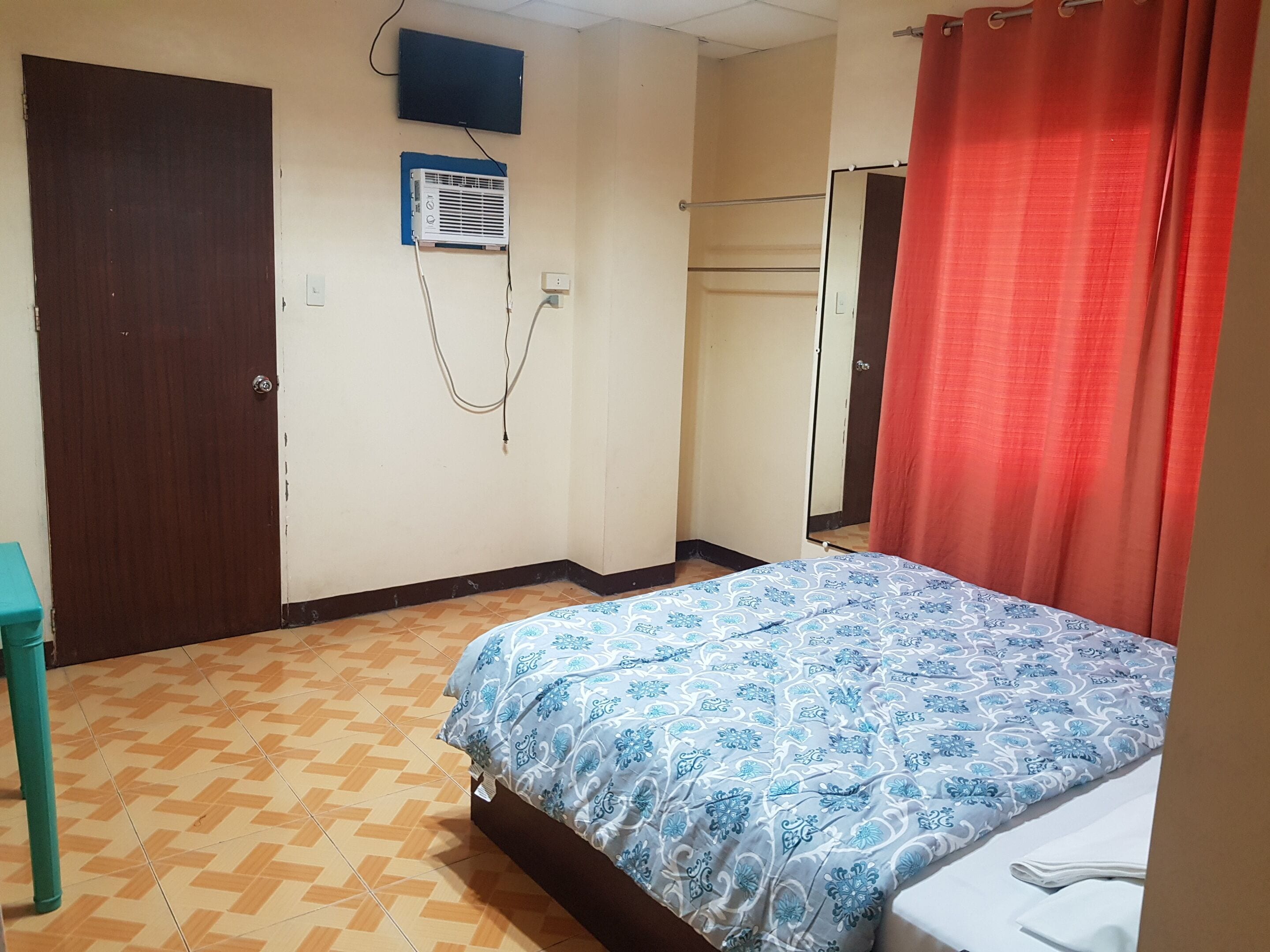 Un dormitorio con una cama, un televisor montado en la pared, un equipo de aire acondicionado y una cortina roja.