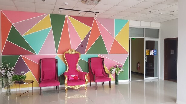 Un espace d’accueil agrémenté d’un mur aux motifs géométriques et aux couleurs vives, de deux fauteuils rouges, d’une chaise à la monture dorée et d’un vase rempli de fleurs.