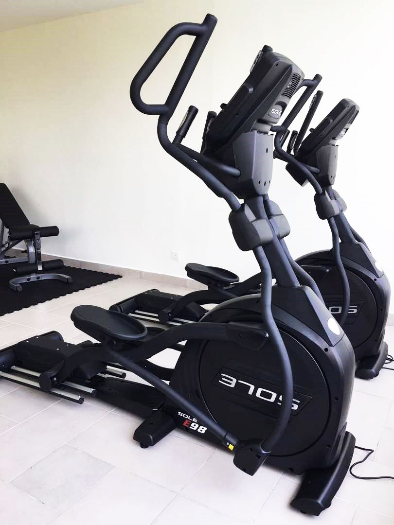Due cyclette ferme in una stanza con pavimento piastrellato e una panca da allenamento nera sullo sfondo.