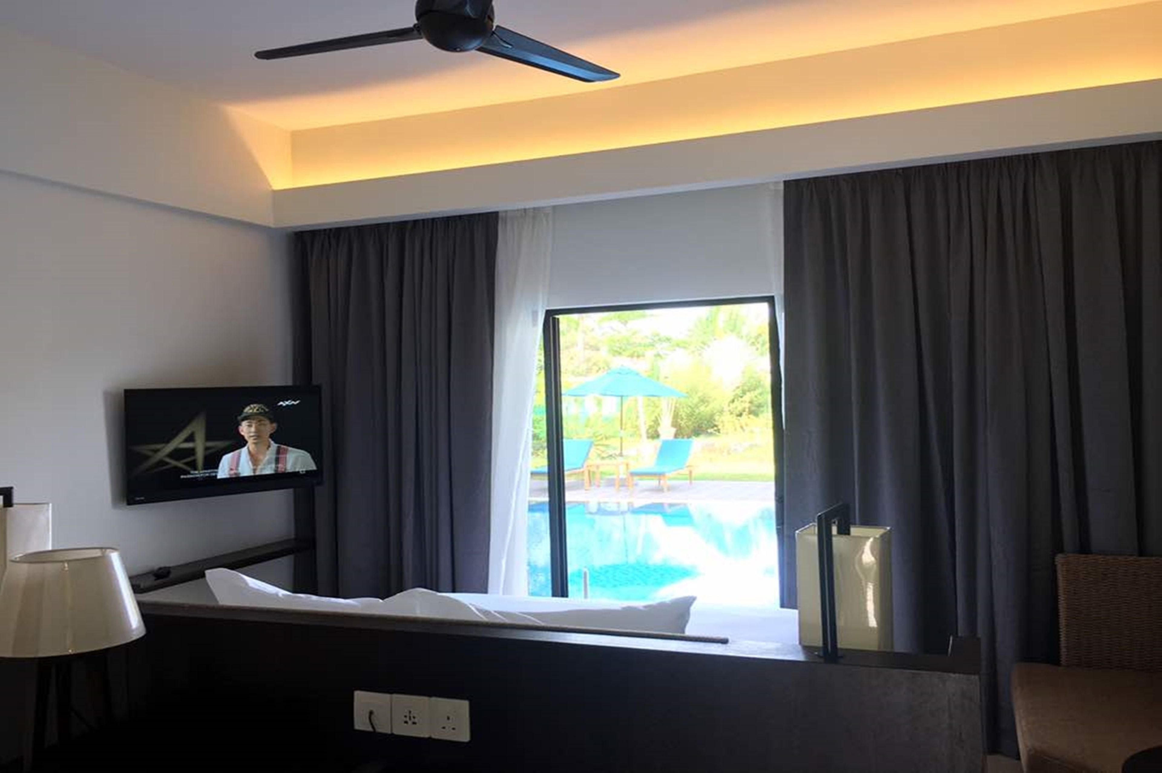 A hotel room with a bed, a television, a ceiling fan, a window with curtains, and a view of an outdoor swimming pool.
