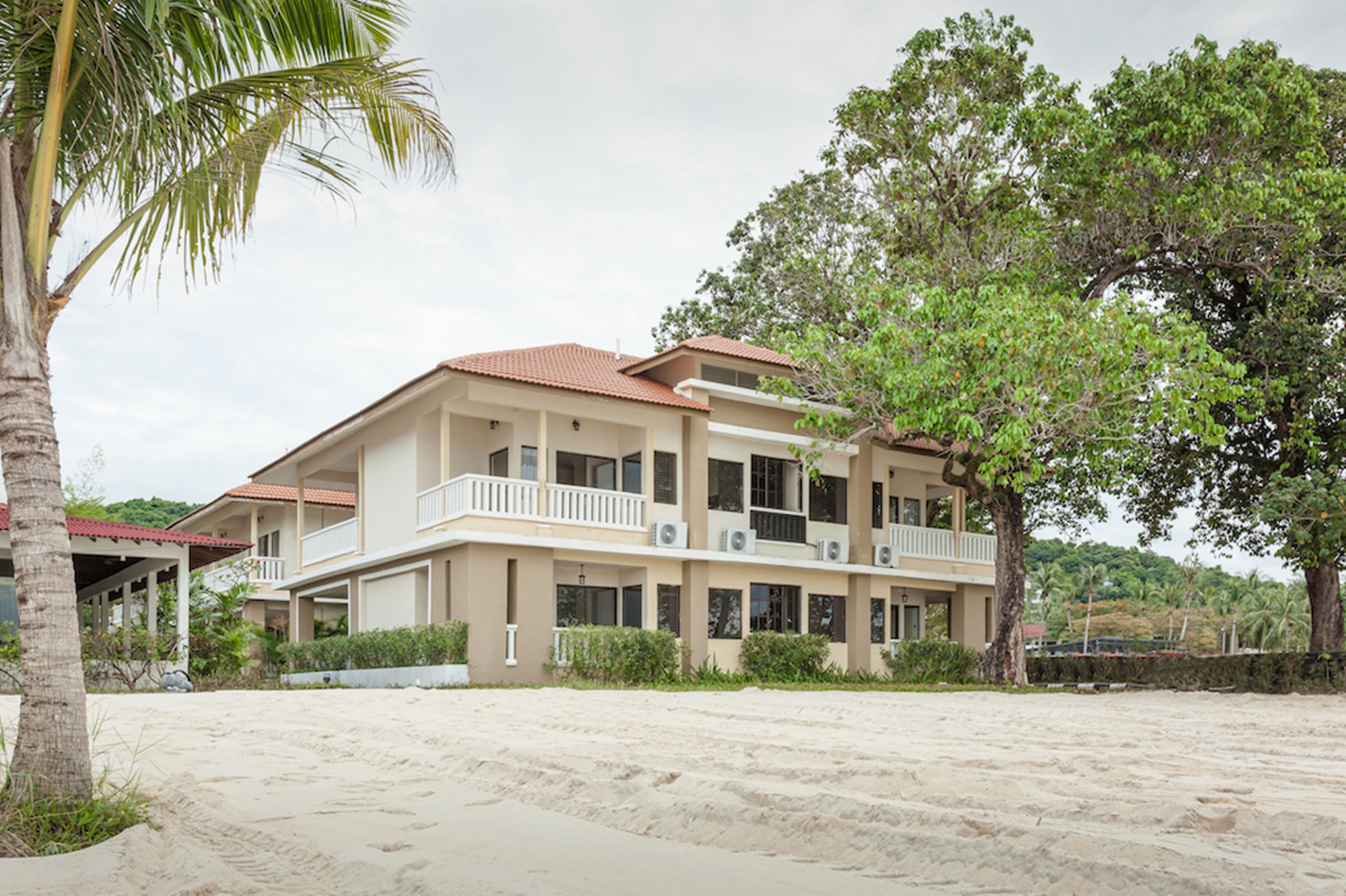A two-story building with a balcony on the second floor, surrounded by greenery and a sandy area.