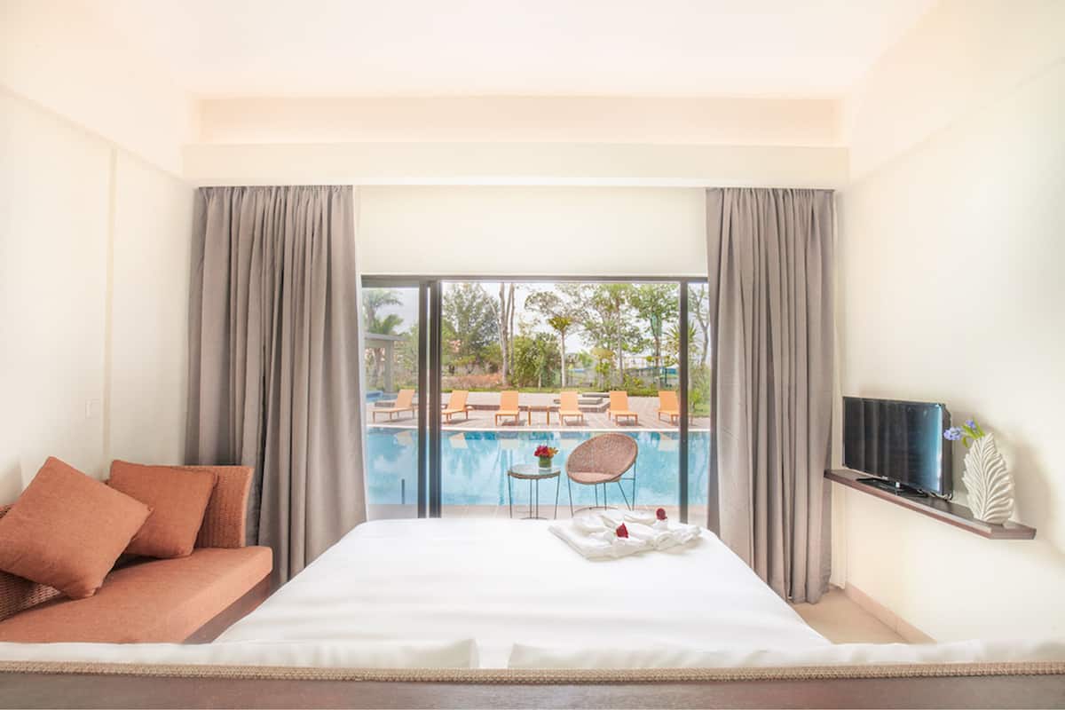 Hotelkamer met een bed, een televisie op een plank, een klein tafeltje met een vaas, en uitzicht op een zwembad en terras.