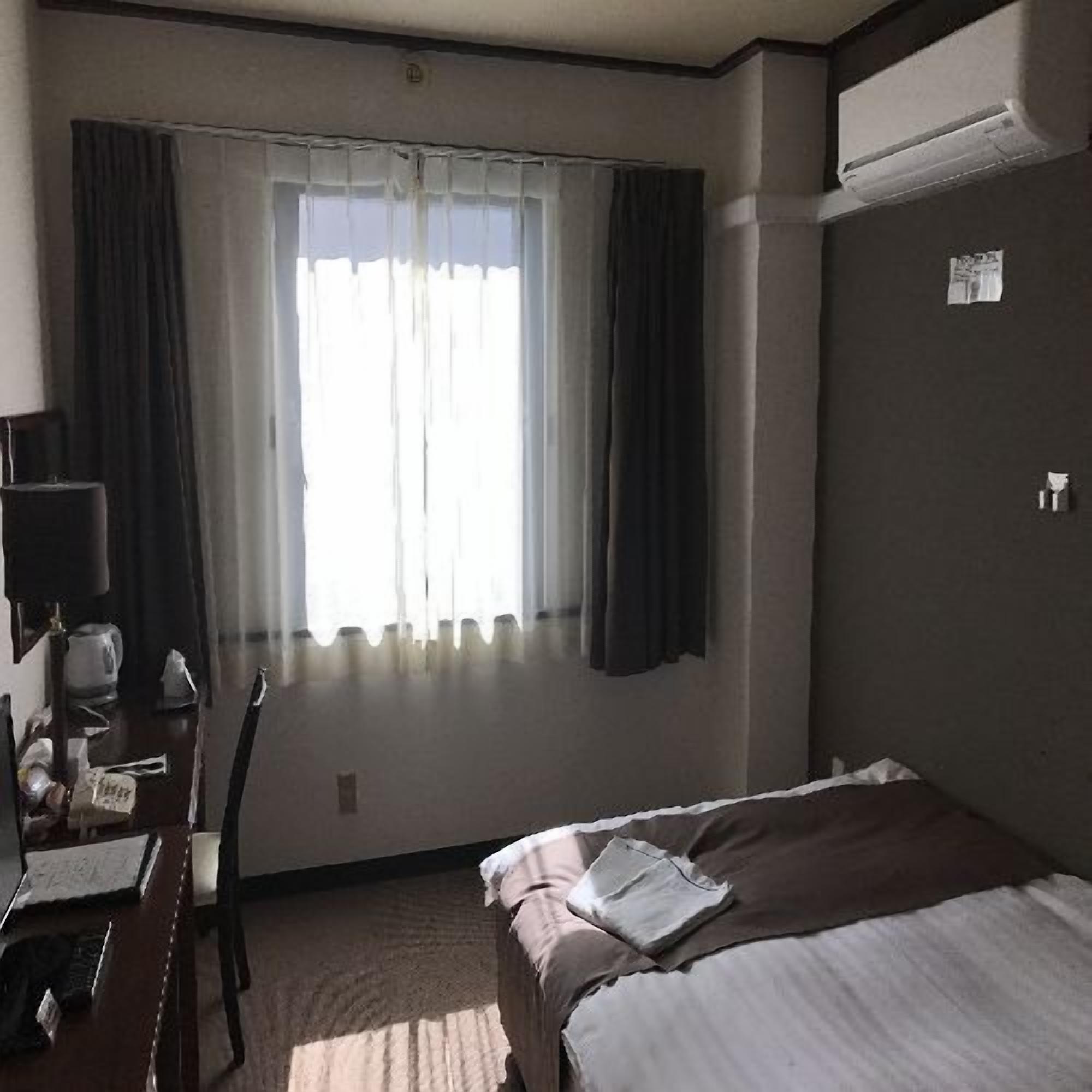 A hotel room with a bed, a desk with a chair, a window with curtains, and an air conditioning unit.