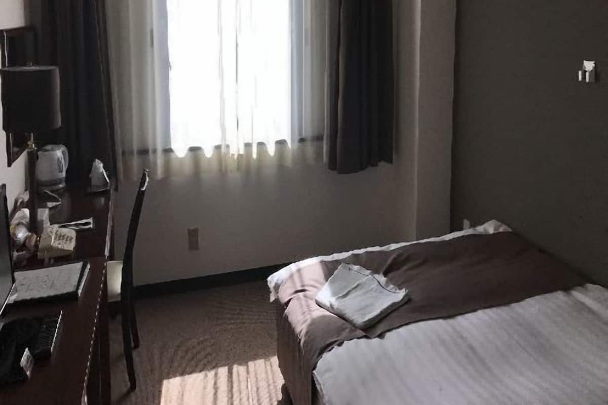 A hotel room with a bed, a desk with a chair, a window with curtains, and an air conditioning unit.