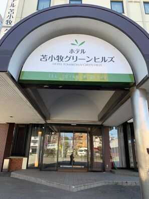 Der Eingang zum Hotel Tomakomai Green Hills mit einem grün-weißen Schild, Glasüren und einer Ziegel-Fassade.