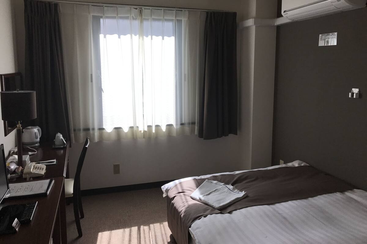 Hotelkamer met een bed, een bureau met telefoon en lamp, een raam met gordijnen en een airconditioning.