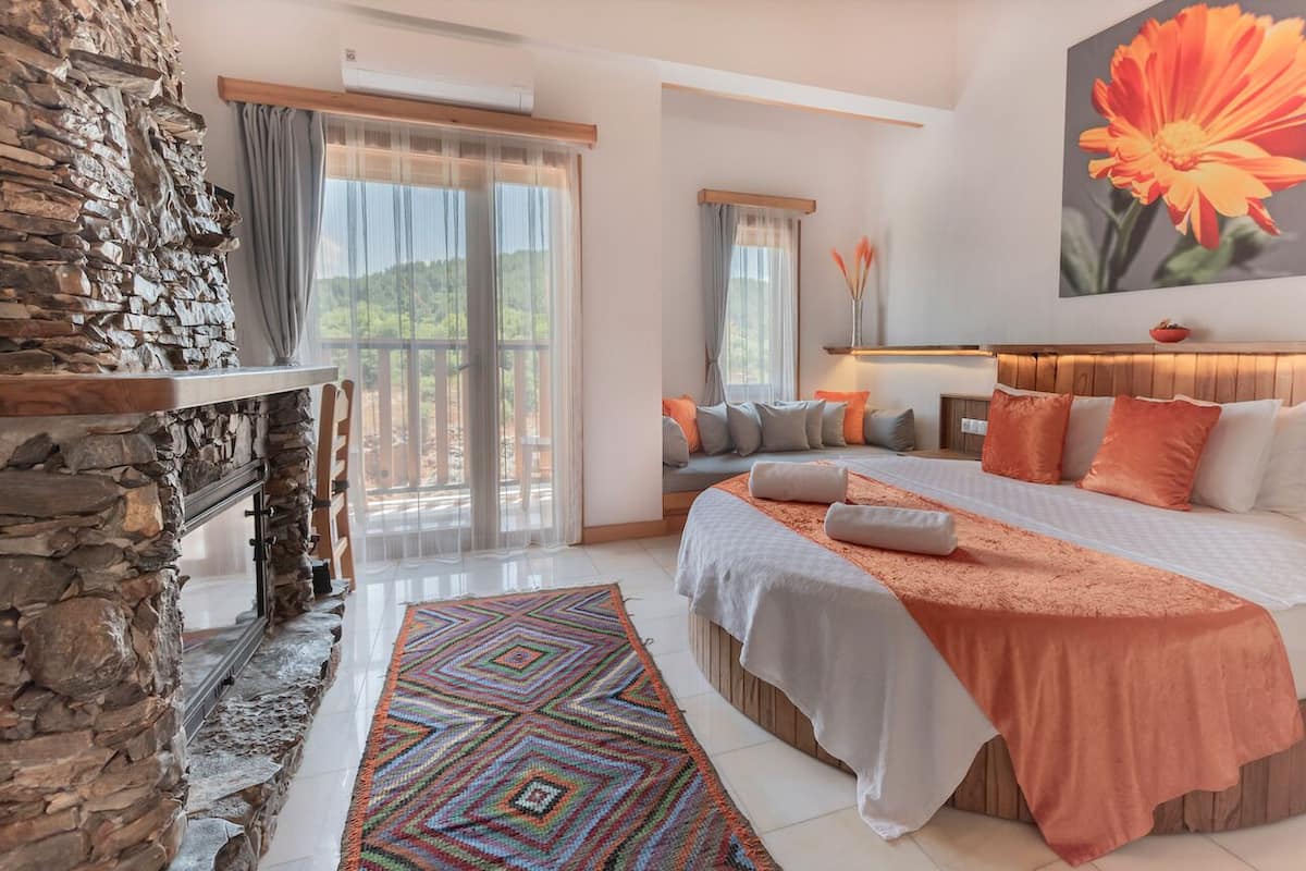 Ein Schlafzimmer mit Kamin aus Stein, einem großen Bett mit orangefarbener Bettwäsche, einem farbenfrohen Teppich und einem großen Fenster mit Vorhängen.