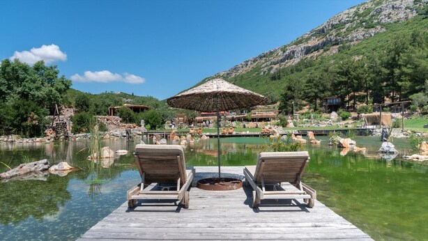 Une terrasse en bois avec deux chaises longues donnant sur une piscine aux eaux cristallines, le tout entouré d’un paysage montagneux et d’une structure à toit de chaume.