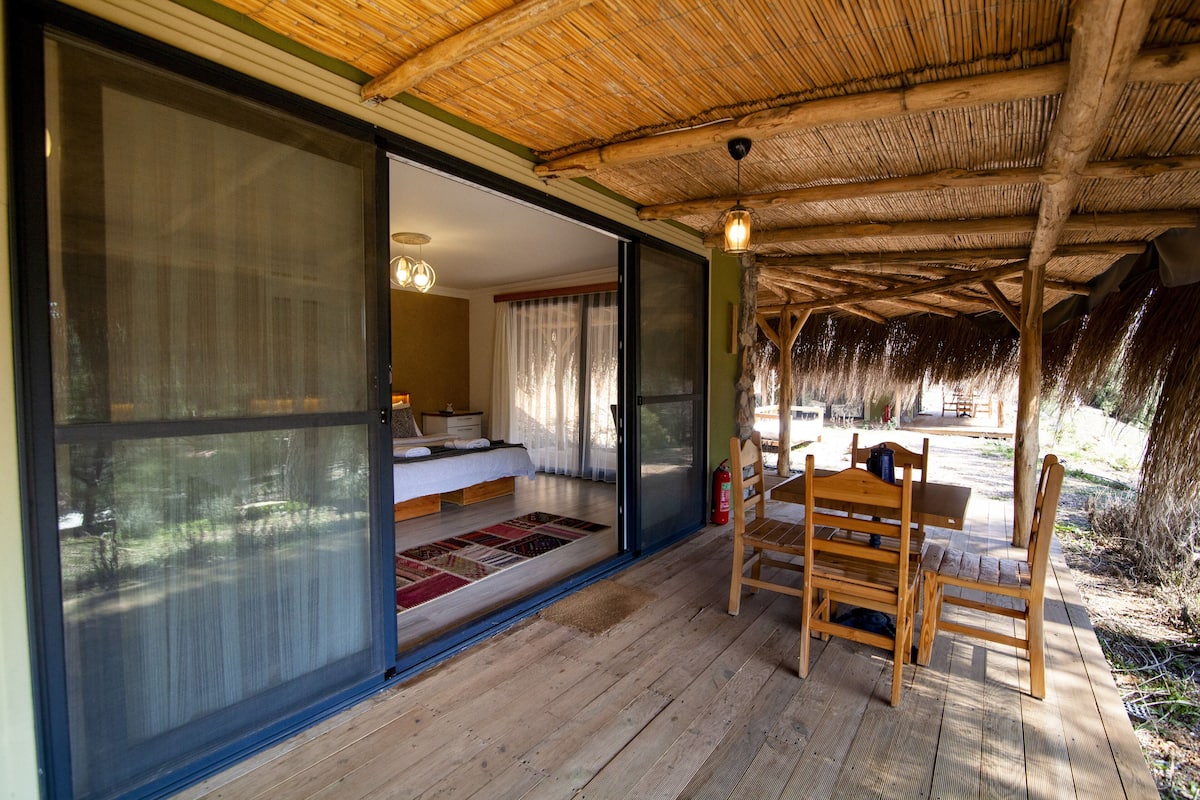 Eine Holzterrasse mit einem Reetdach, eine Glasschiebetür zu einem Schlafzimmer und ein Essbereich mit Holzzmöbeln.