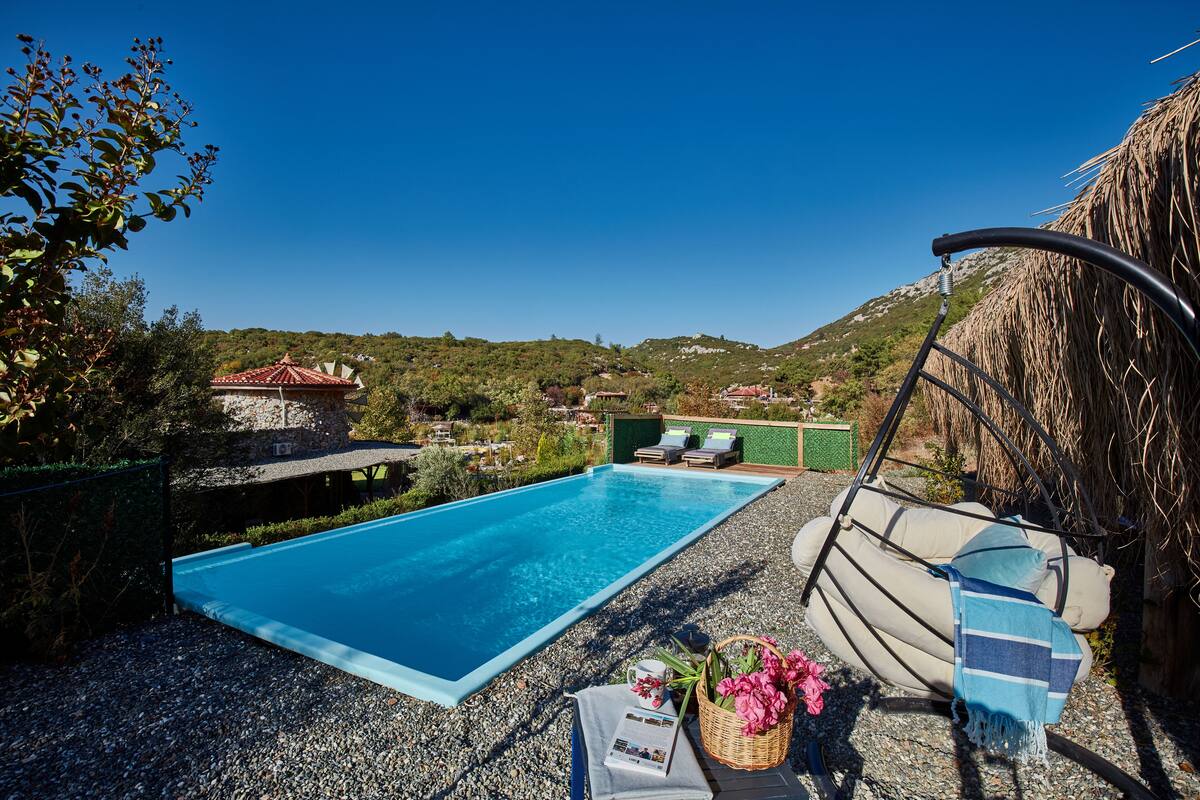 Ein klares, blaues Schwimmbecken mit Liegestühlen und einer Hängematte, umgeben von einer felsigen Landschaft und einer entfernten Ansicht von Häusern.