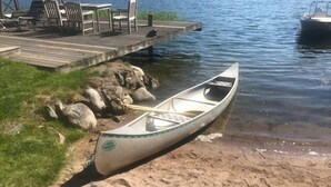 Un canoë est amarré à un quai, près d’une plage de sable, avec une zone herbeuse et des rochers à proximité.