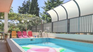 A swimming pool with a tiled edge, a covered area with chairs, and a fenced garden.