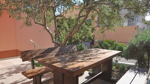 An outdoor wooden table and benches in a shaded area with a building in the background.