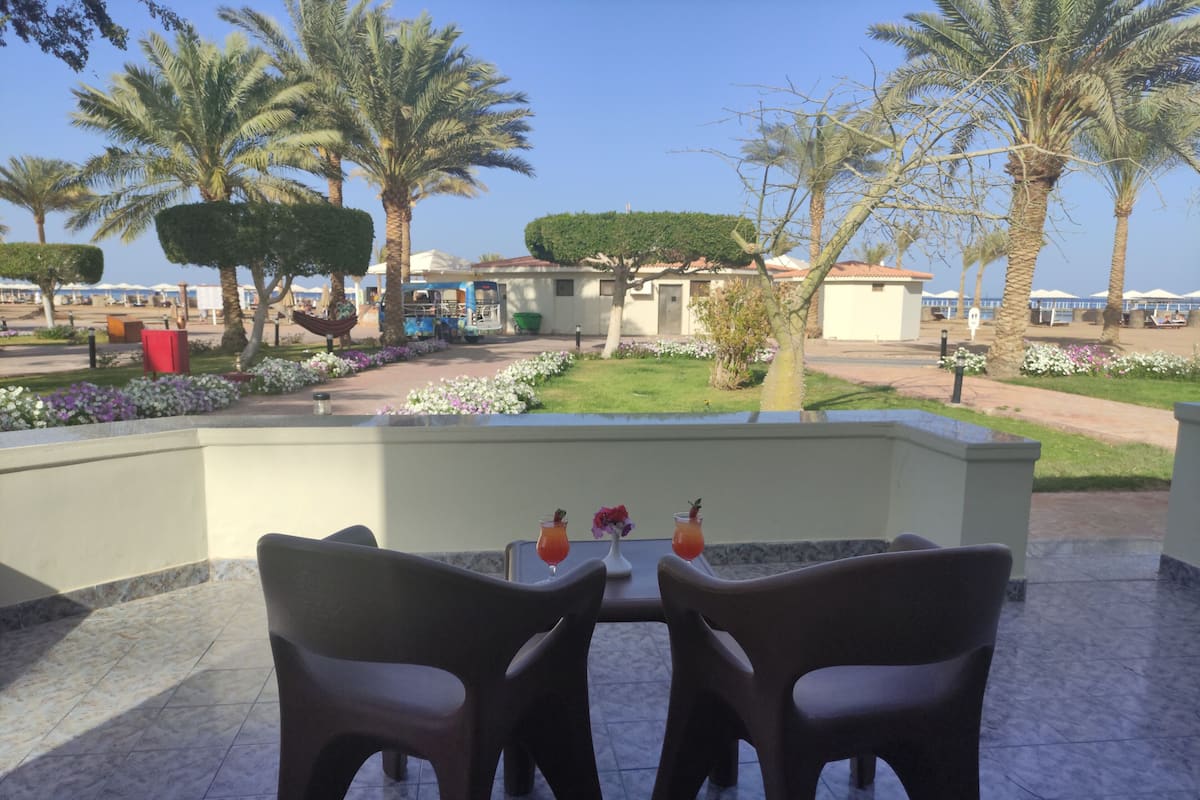 A patio with two chairs and a table, overlooking a landscaped garden with palm trees and a view of a building with a red roof.
