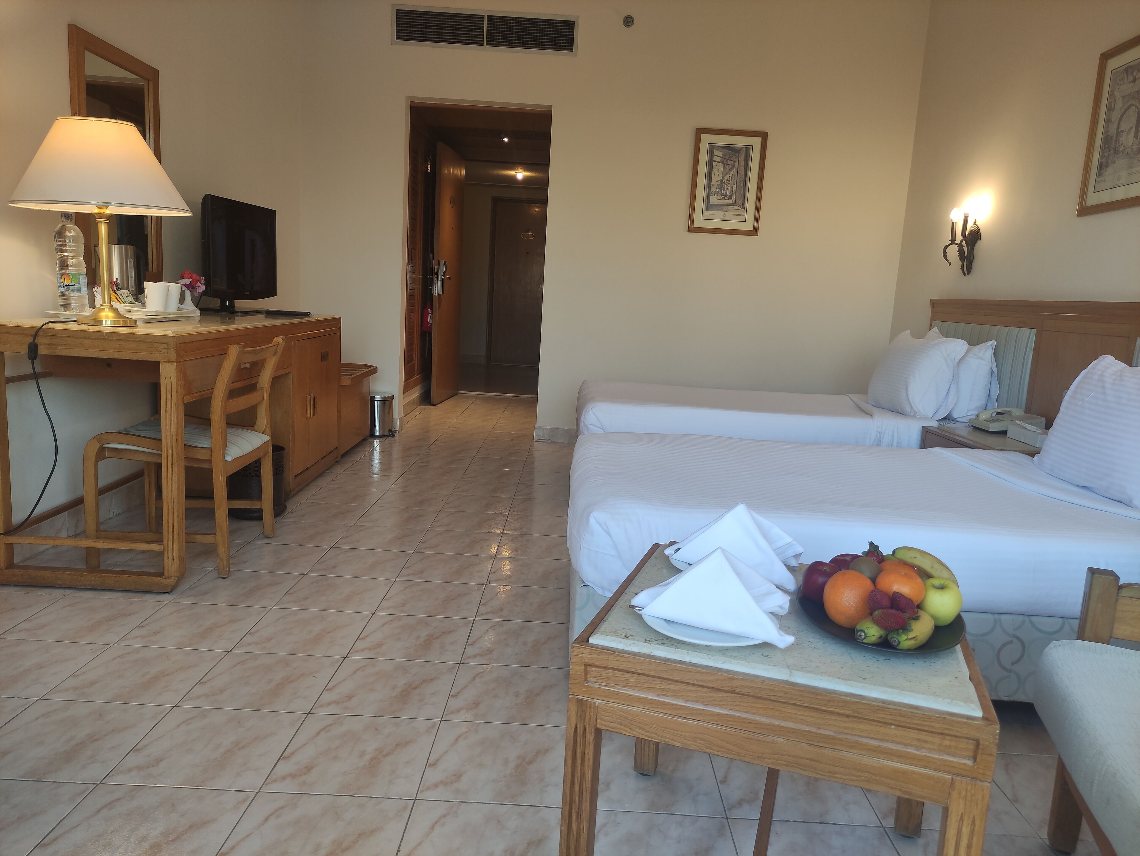 Ein Hotelzimmer mit Schreibtisch, zwei Betten, einem kleinen Tisch mit Obst und einer Lampe.
