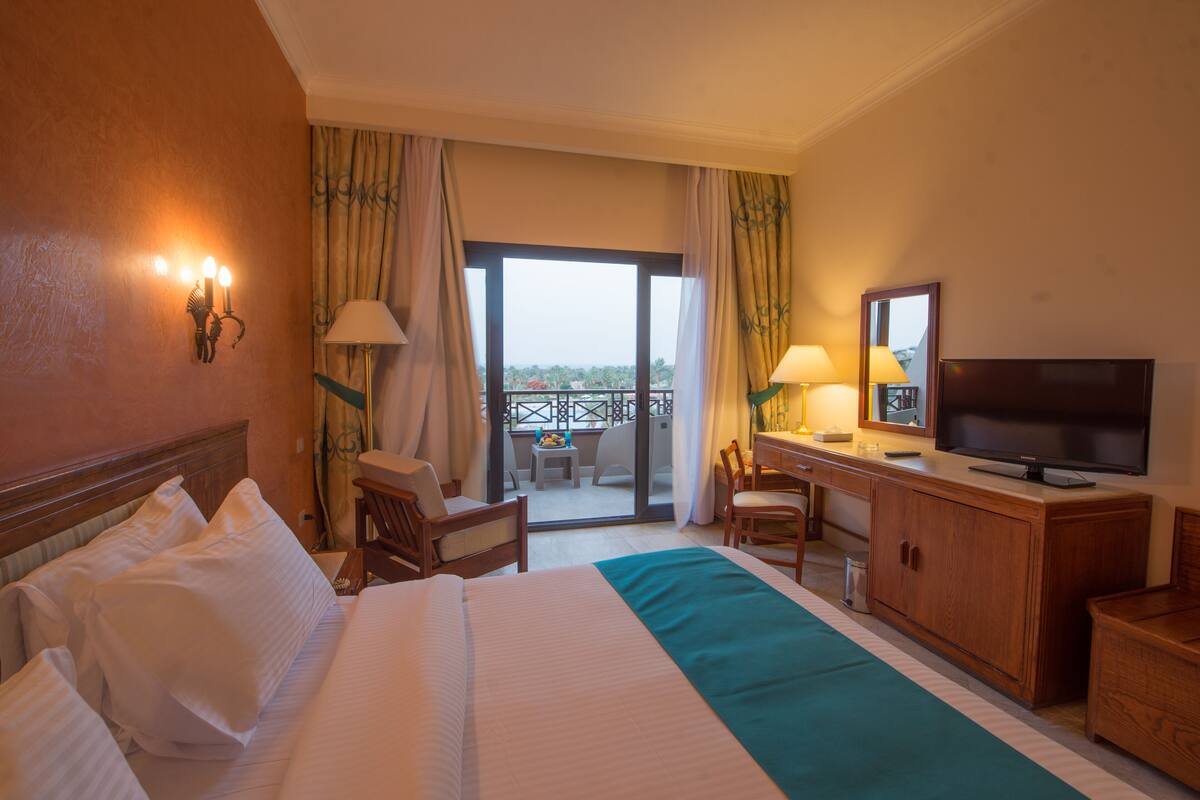A hotel room with a bed, a desk with a TV, a chair, a balcony with a view, and a window with curtains.
