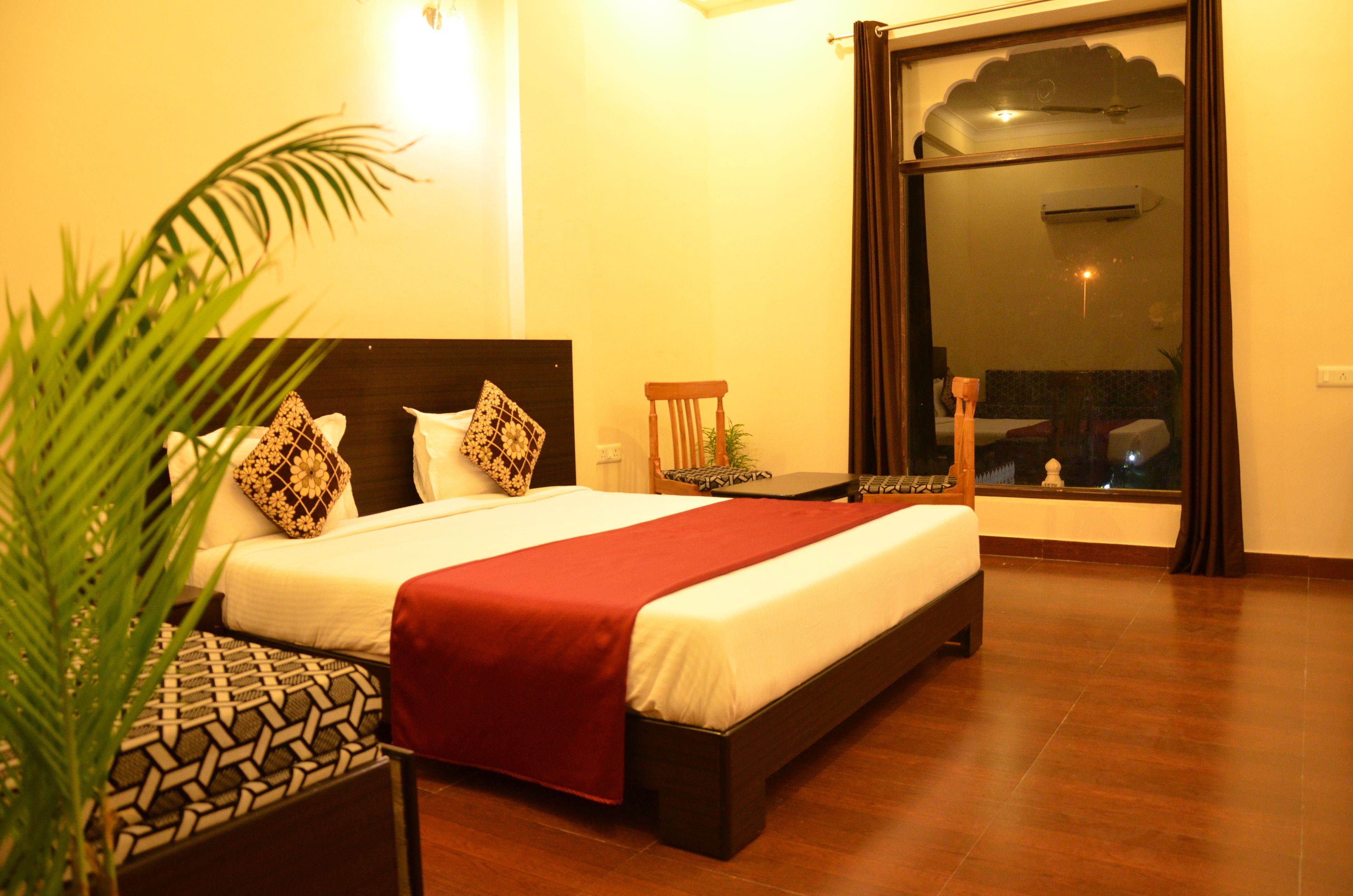 Habitación de hotel con una cama grande, una manta roja, una mesita de noche de madera, una planta en maceta y vistas al exterior a través de una puerta corredera.