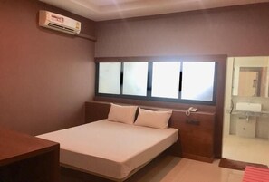 A bedroom with a bed, a window, an air conditioning unit, and a bathroom visible through an open door.