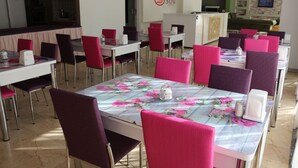 A dining area with tables and chairs, a kitchen in the background, and a logo on the wall. - Suw Hotel (Atakum)