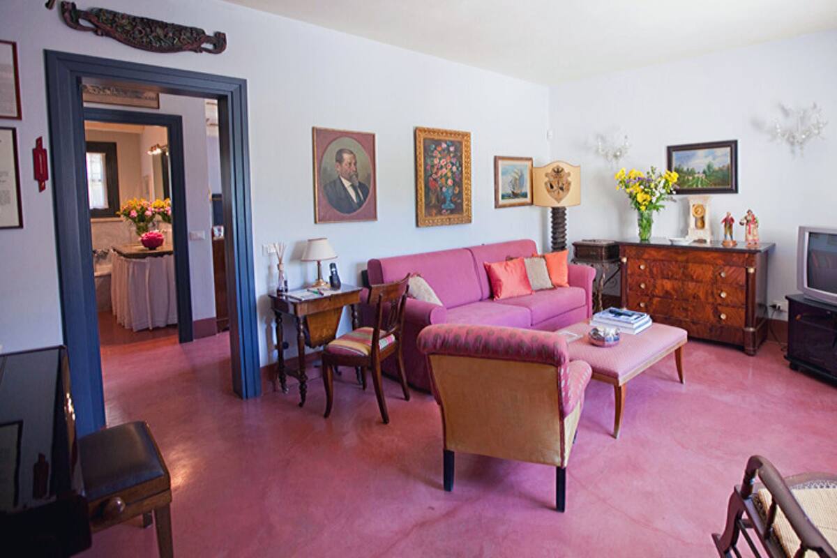 A living room with a pink sofa, wooden chairs, a coffee table, and a television. There are framed pictures on the wall and a cabinet with decorative items.