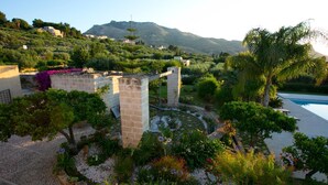 Un jardín con piscina, estructuras de piedra y montañas al fondo.