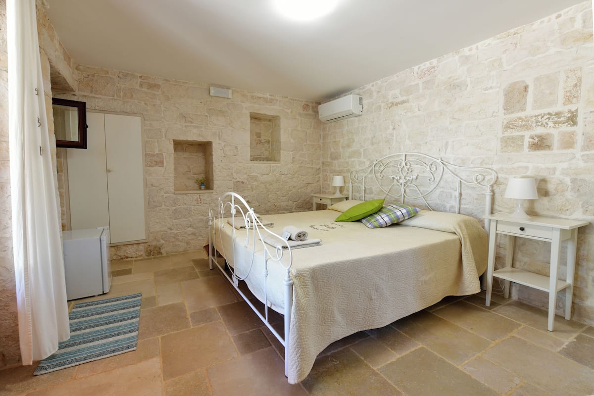 Een slaapkamer met een stenen muur, een bed met wit metalen frame, een nachtkastje met een lamp en een klein wit kastje.