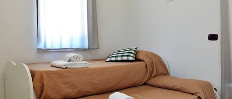Une petite pièce comprenant un lit, une fenêtre avec des rideaux, un luminaire fixé au mur et une prise de courant.