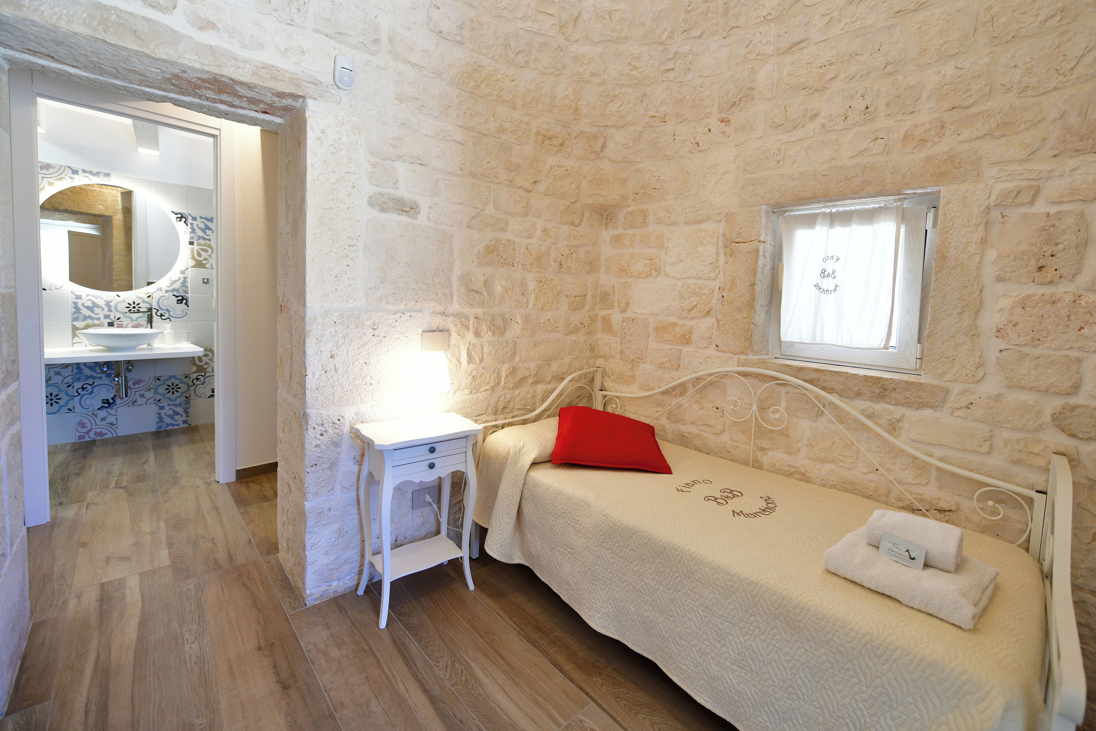 A bedroom with a stone wall, a bed with a red pillow, a bedside table, and a window with a curtain.
