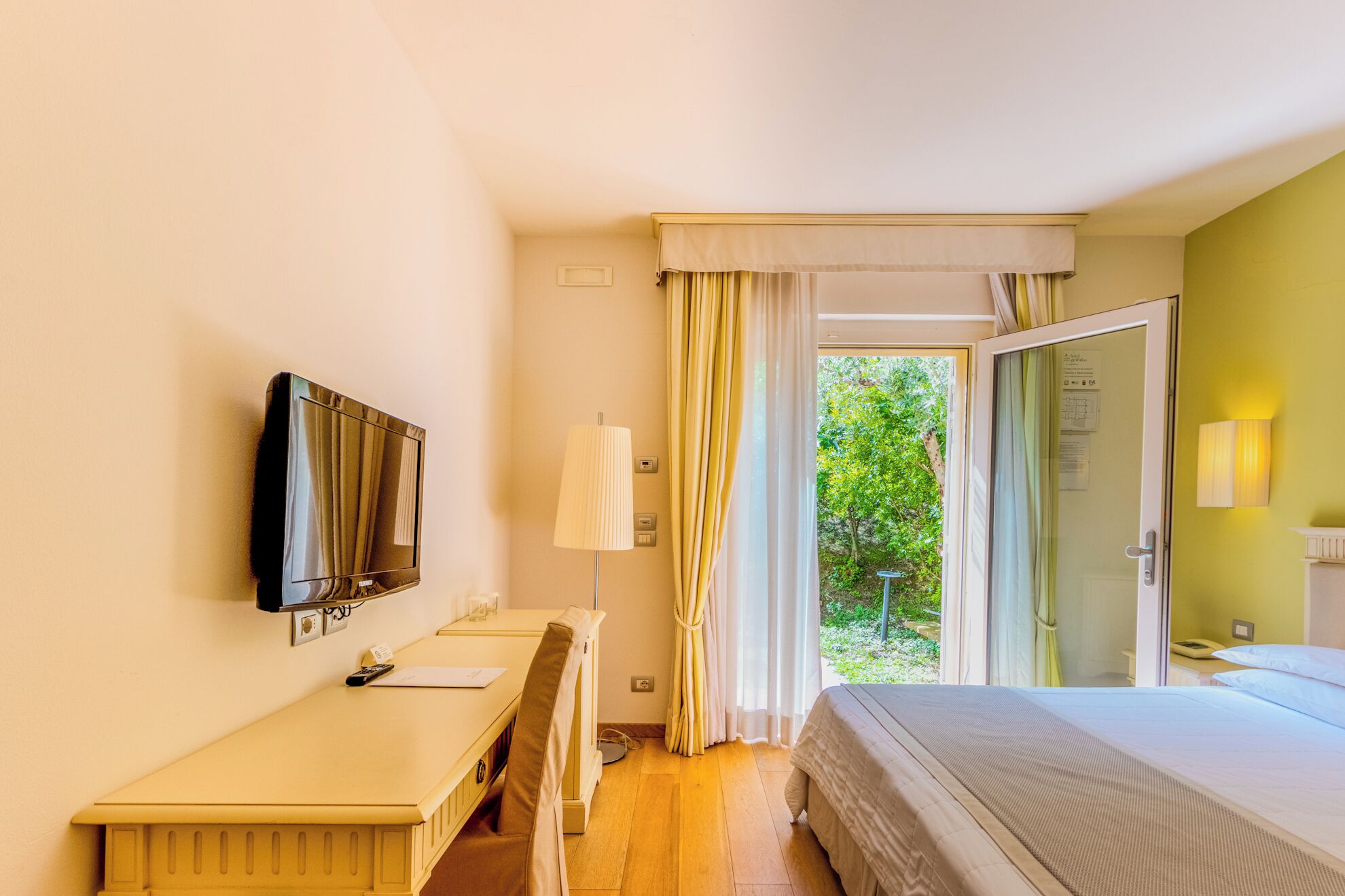 A hotel room with a bed, a desk, a television, a lamp, and a sliding glass door leading to a garden.