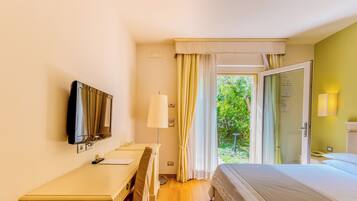 Habitación de hotel con cama, escritorio, televisor, lámpara y puerta corrediza de vidrio que da a un jardín.
